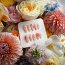 Handmade floral press-on nails with 3D sculpted pink flowers and crystal accents on an almond shape, displayed on a Joyee Nails card surrounded by fresh dahlias, roses, and wildflowers