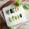 Handmade green nail designs by Joyee Nails featuring 3D frog art, dark green leaf accents, pastel floral patterns, and matcha green ombre on almond-shaped press-on nails — cute and whimsical green nail ideas for spring.