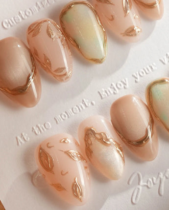 Close-up of Earthy glow press-on nails with nude peach color, gold foil and leaf accents